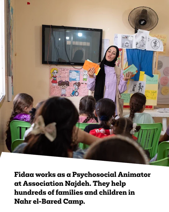 ‘Fidaa works as a Psychosocial Animator at Association Najdeh. They help hundreds of families and children in Nahr el-Bared Camp.’
Fidaa is stood in a classroom, teaching children who are sat down. 
