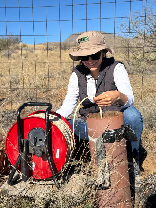 A scientist sits, smiling, while she slowly lowers the tape of a water level sounder into a well.