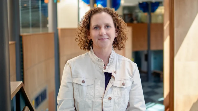 A person with curly hair wearing a light jacket and patterned blouse, smiling in a modern office setting.