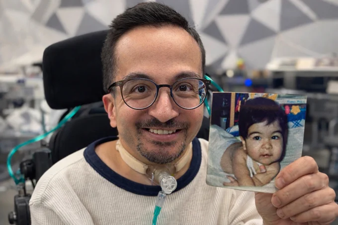 AI-generated image of an adult man smiling in a wheelchair, holding a small printed photo of himself as a baby, with medical equipment visible in the background.