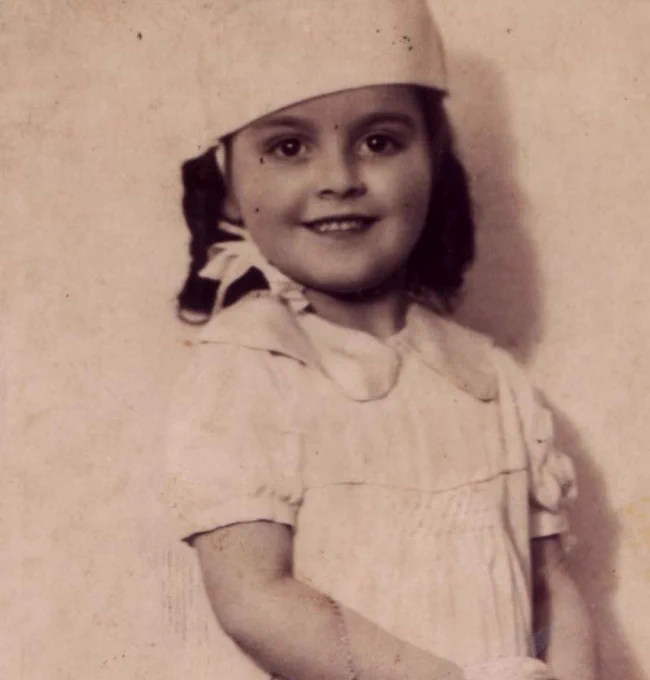 Vintage photograph of a smiling  wearing a cap and a dress.
girl