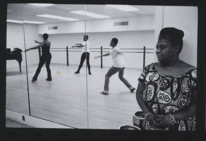 Black and white image of a woman wearing a dress with her hair in a bun, watching three dancers in a dance studio. The dancers are visible in a mirror behind the woman.