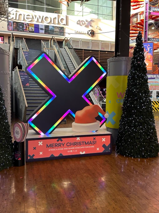 Large X-shaped sculpture with neon lights displayed by two Christmas trees at Xscape Yorkshire.