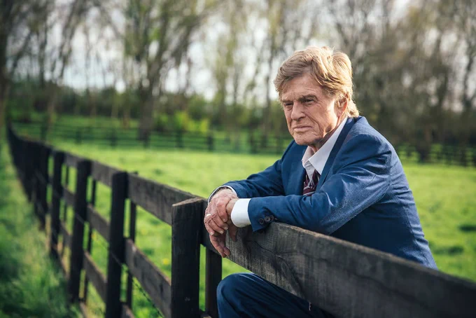 Robert Redford leans over a wooden fence.
