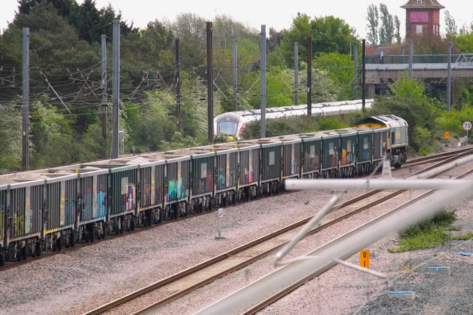 Freight train composed of multiple cars loaded with cargo, moving along tracks in a rail yard with trees and a control tower in the background.