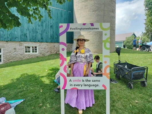 Person dressed in traditional attire posing behind a colorful frame with the message 'A smile is the same in every language' at an outdoor event.