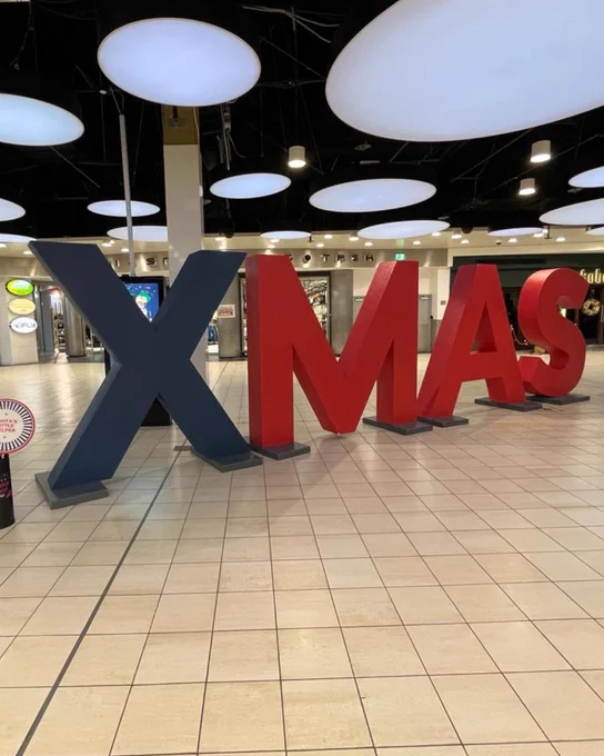 Large "XMAS" letters in red and blue displayed in a public indoor space with white, cloud-shaped lights hanging from the ceiling.