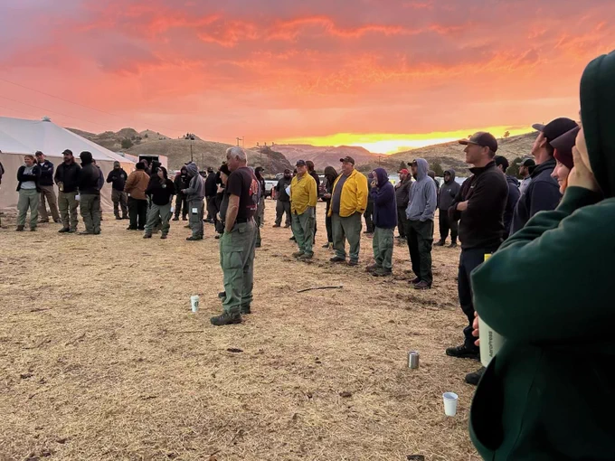 A few dozen firefighters gather for an outdoor meeting with a gorgeous pink sunrise in the background. Firefighters gather for the morning briefing on the Black Rock Fire in central Oregon.