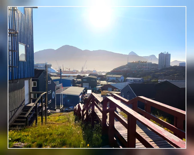 Blick auf den Hafen und das Industriegebiet von Nuuk, in Hintergrund sind Berge, Blätter Himmel und die Sonne.
Im Vordergrund ist eine Treppe, denn wie auch in allen anderen Orten von Grönland geht es in der Stadt bergauf und -ab.