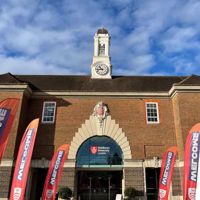 Front view of the entrance to Middlesex University in London, featuring a classical building with a clock tower, under a clear blue sky. Red welcome banners flank the doorway.