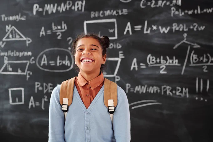 Smiling girl with backpack in a classroom, standing in front of a chalkboard with math formulas.