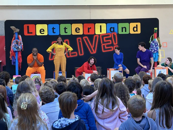 6 actors dressed in various monotone colors stand in the gym in front of a backdrop that reads "letterland alive!" they are in various poses and have an audience of students in front of them