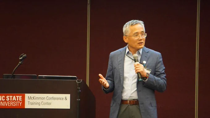 Dr. Paul Liu stands at the front of a room holding a microphone, presenting