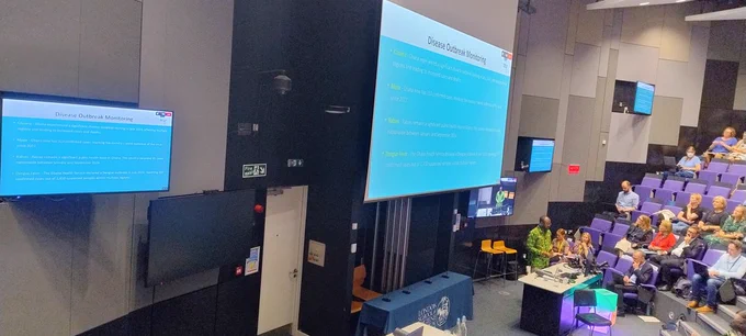 Rabiu Alhassan, Managing Editor of GhanaFact and Director of FactSpace West Africa, speaking at the LSHTM Health Misinformation UNPACKED event. Display screens showing slides and a video screen can be seen behind Rabiu, along with attendees at the event.

