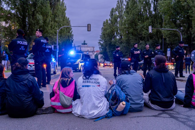 Aktivist:innen sitzen auf einer Straße, Polizei steht um sie herum