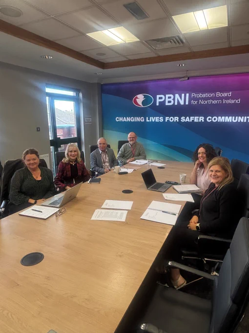 Photo of four women and two men sitting at a table in front of a large wall mounted Probation Board for Northern Ireland logo.