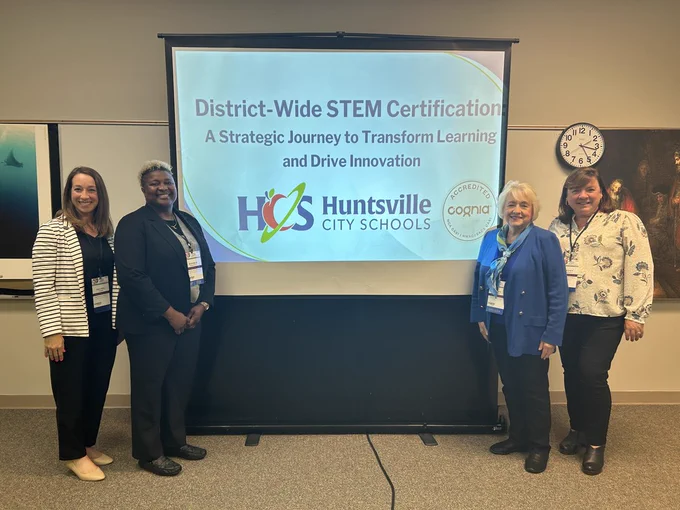 Rachel Evans, Bonnie Garrett, Cathy McNeal, and Rebecca Campbell at the Cognia Conference, Powerpoint slide behind them which reads District-Wide STEM Certification, a Strategic Journey to Transform Learning & Drive Innovation