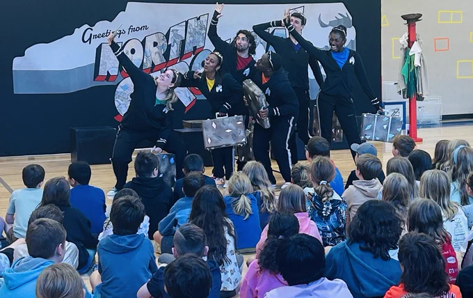 actors in black outfits have their hands up in the air and stand in front of a backdrop that reads "greetings from north carolina"