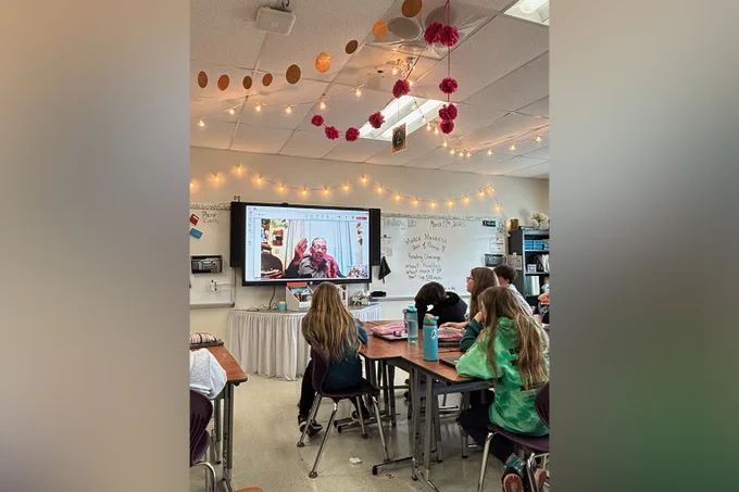 A classroom of kids watches a projection screen featuring a man talking to them through a video calling platform