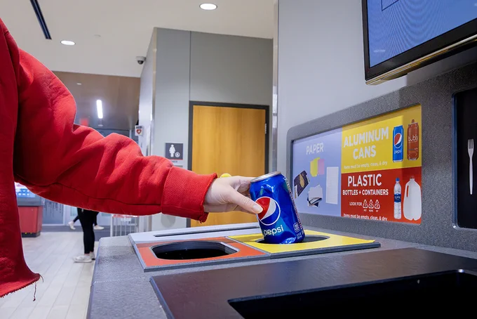 A person in a red jacket drops a Pepsi can into a bin at an All in the Hall recycling station. (Kristen Labadie/University Communication and Marketing)