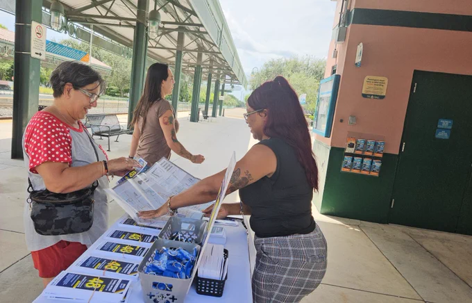 A woman with glasses and long hair is standing behind a table at a transit station, handing a brochure to an older woman in a red shirt with white polka dots. Another woman walks past in the background. The table displays route brochures, pens, and small giveaway items.