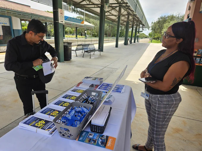 A man in a black shirt is standing at an outdoor table with informational brochures and giveaway items, filling out a form. Across from him, a woman with glasses and long hair listens attentively. The setting is a transit station platform with benches and a covered walkway.