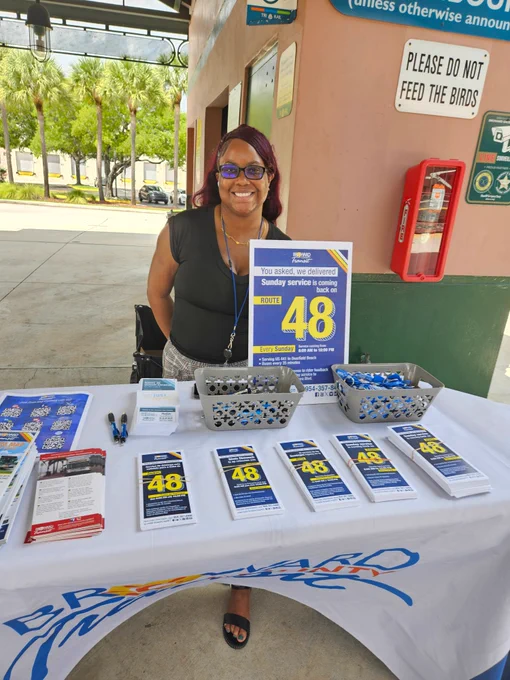 A smiling woman with glasses and long hair stands behind a table covered with Broward County Transit materials. A large sign on the table reads “Route 48 – Sunday service is coming back.” The table is neatly arranged with brochures, pens, and promotional items, with a transit station wall and palm trees visible in the background.