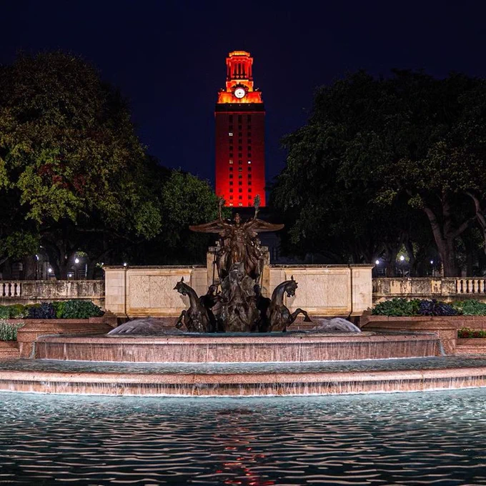 The image shows the University of Texas Tower illuminated in burnt orange behind the Littlefield Fountain, captured at night.