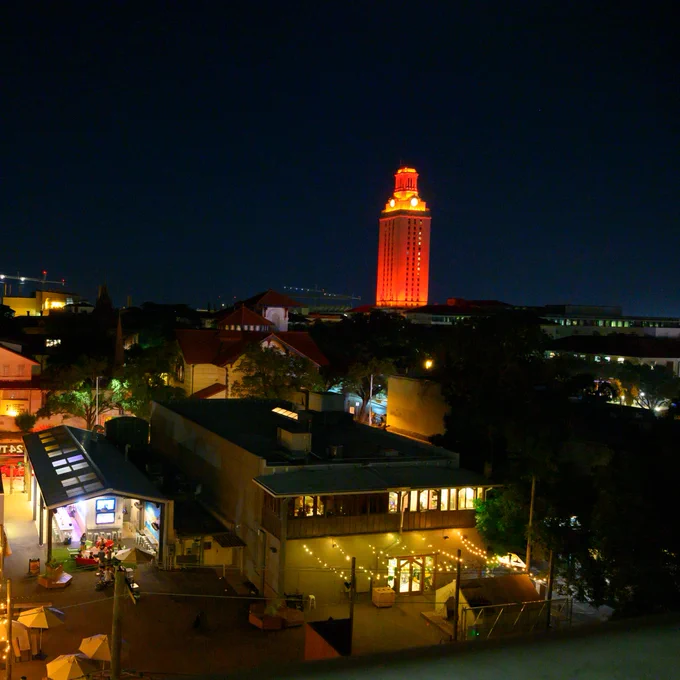 Nighttime view of Austin featuring illuminated buildings and streets, with the iconic UT Tower lit up in orange in the background.