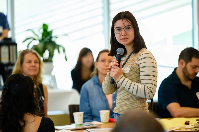 A Harvard College student speaks at the event, holding a microphone and standing. Seated participants behind them look on.