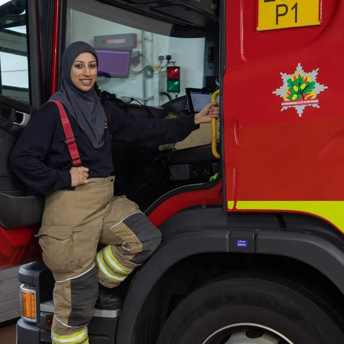 A woman poses next to a firetruck, holding its handle and foot up. She smiles to camera, wearing her uniform and hijab.