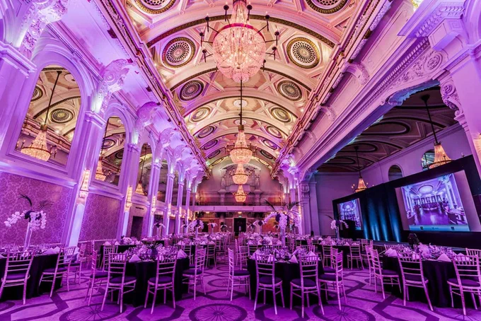 Great Hall at De Vere Grand Connaught Rooms in London, a large baroque mirrored hall with crystal chandeliers under a high ornate dome, lit in amber and violet light and set for a formal dinner.