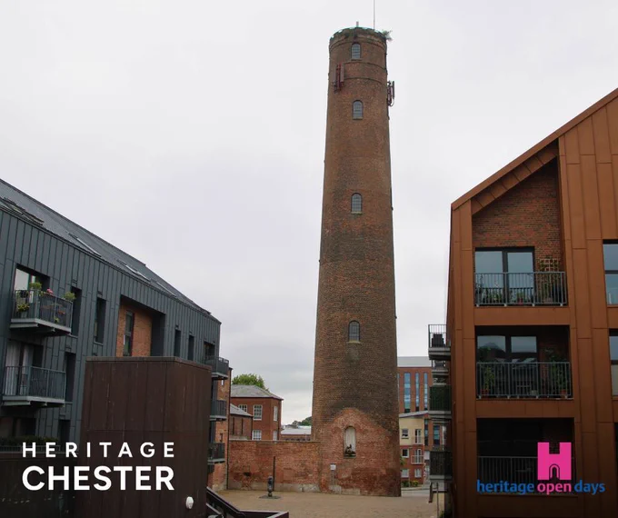 A photograph of Chester Shot Tower, a tall cylindrical brick tower constructed from red sandstone, standing prominently in the center of a modern residential development. The historic industrial tower features small windows at regular intervals up its height and appears to be several stories tall. It is surrounded by contemporary apartment buildings with modern architecture - including buildings with grey metal cladding and orange/brown facades with balconies. The contrast between the Victorian industrial heritage and modern housing development is clearly visible. The sky appears overcast. In the bottom left corner is white text reading "HERITAGE CHESTER" and in the bottom right is the Heritage Open Days logo featuring a pink castle icon with blue and pink text.