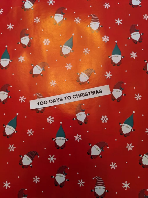 100 days to Christmas written on Christmas wrapping paper covered in snowflakes and Father Christmas gnomes