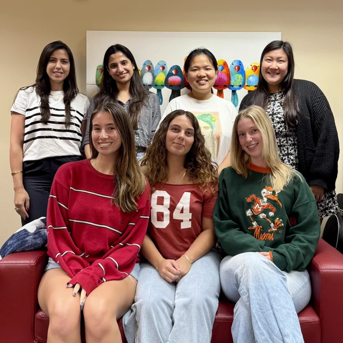 Seven smiling women in two rows on red couch with colorful bird painting in the background.