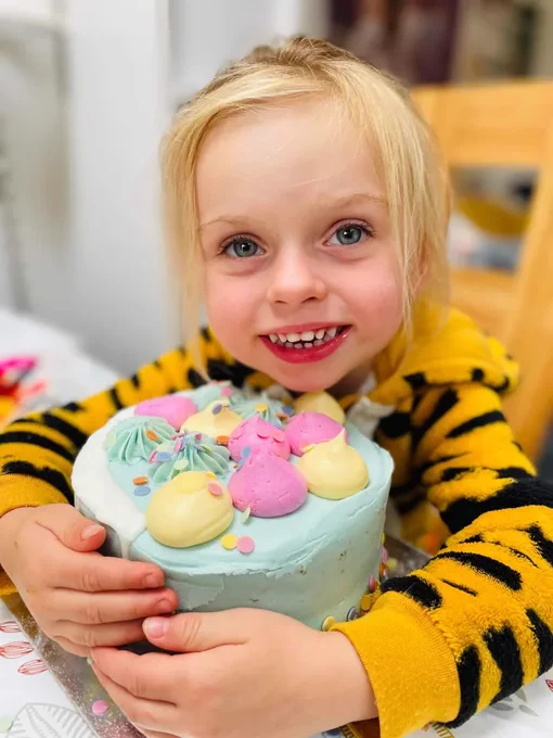 A child with a big smile holding a pastel-colored birthday cake topped with decorative icing.