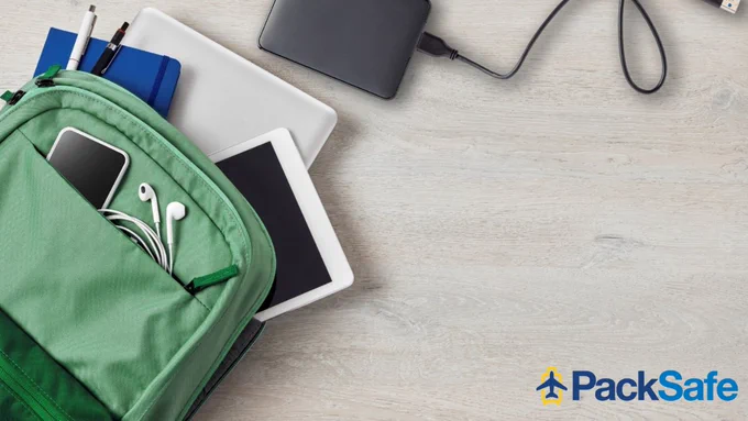 Green backpack with electronic devices like tablets and a smartphone, headphones, and notebooks on a wooden surface. PackSafe logo visible on the right.