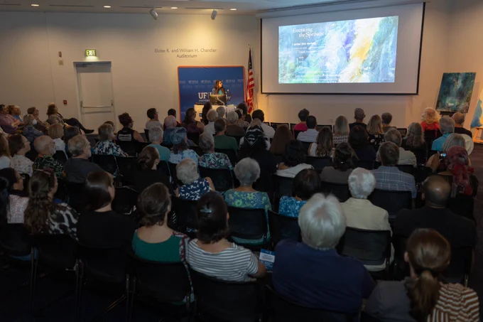 A room full of people watching a lecture. A speaker is at the front of the room and a projector screen.