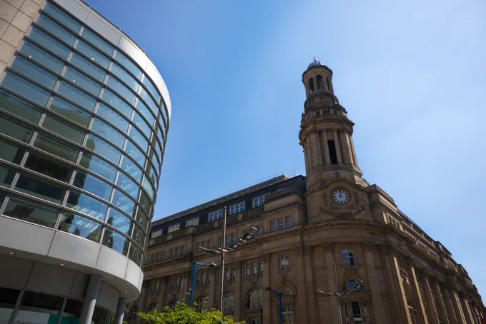 Manchester Corn Exchange building from a low angle against a bright blue sky and juxtaposed with a modern, glass-fronted building