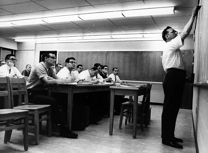Black and white image of a teacher writing on a blackboard while a class looks on.