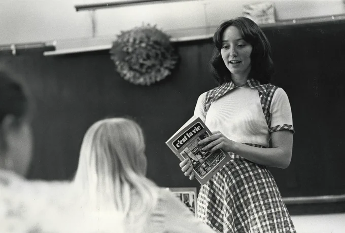 Black and white image of a woman wearing a plaid skirt and white top standing in front of a blackboard holding a book titled "c'est la vie"