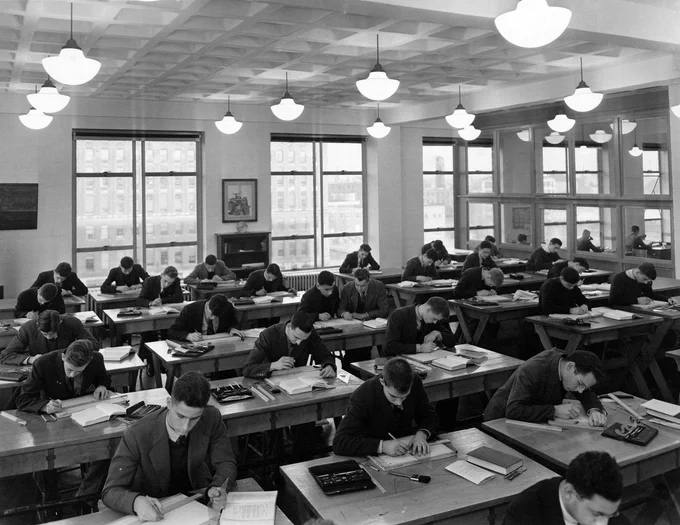Black and white image of a classroom of men bent over desks, working with notebooks, rulers, books, and other materials. The windows behind them show views of the city