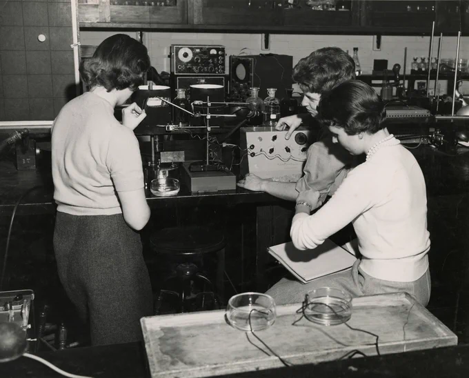 Black and white image of three women working on a machine in a lab
