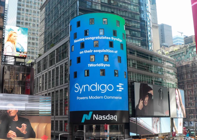 A daytime photograph of the large, cylindrical Nasdaq billboard in a bustling Times Square. The billboard's screen displays a congratulatory message, which reads: "Nasdaq congratulates Syndigo on their acquisition of 1WorldSync." Below this message are the Syndigo and Nasdaq logos, along with Syndigo's tagline, "Powers Modern Commerce."