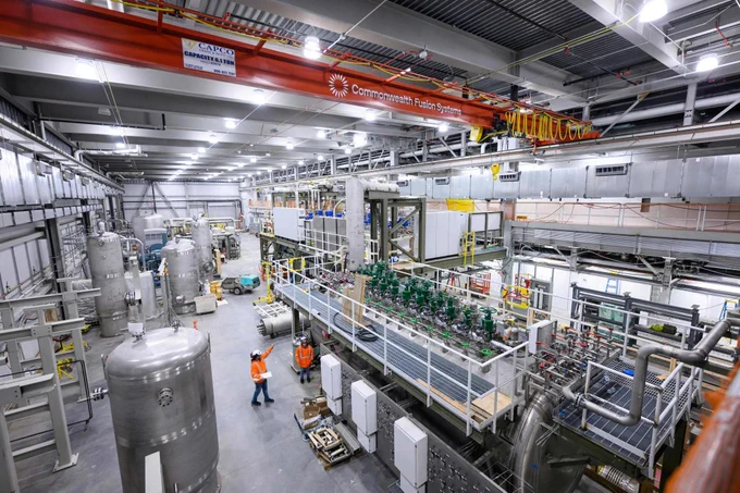 A large industrial room packed with high-tech cooling equipment, green valves, silver tanks, with an orange crane overhead.
