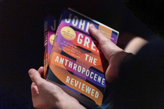 A pair of hands holds two copies of John Green's book "The Anthropocene Reviewed."