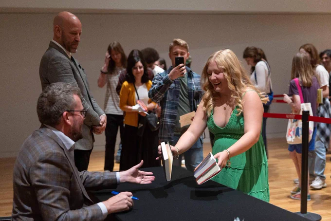 An attendee hands John Green a stack of books to be signed. More attendees wait in line in the background.
