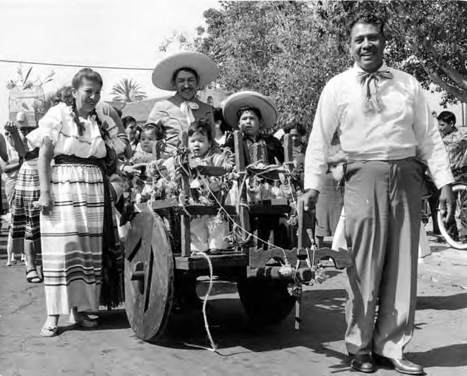 A black-and-white photo of a festive parade scene with a woman in traditional dress, a man in a wide sombrero, and children riding in a decorated wooden cart with large wheels, surrounded by other people.