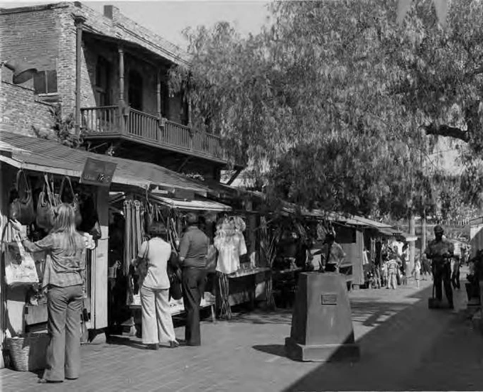 A black-and-white photo of shoppers browsing outdoor vendor stalls at Olvera Street lined with clothing and goods, set against a historic two-story brick building with a wooden balcony.