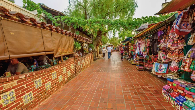 A vibrant outdoor walkway on Olvera Street in Los Angeles, lined with red brick tiles. On the left, El Paseo Inn restaurant features colorful tiled walls and shaded seating, while on the right, market stalls display clothing, souvenirs, and crafts under bright awnings. People stroll down the path beneath lush green trees.